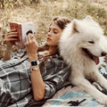 Woman reading a book with a white dog lying next to her on a blanket in a field.