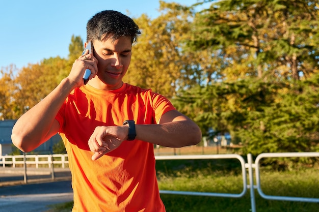 Man in orange shirt using phone and checking watch outdoors with trees in background